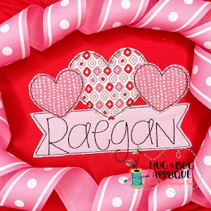 以下が含まれることがあります： 赤い布地に、ピンクのハートが3つと、「Raegan」という名前が刺繍されたバナーが縫い付けられています。ハートは、水玉模様や正方形など、さまざまなパターンで飾られています。バナーはピンクと白のストライプ模様です。