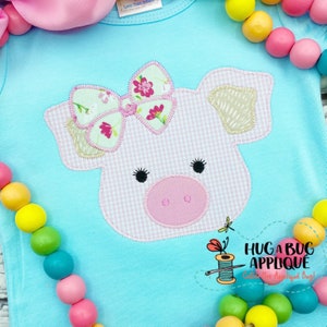 May include: A pink pig applique with a floral bow on a light blue shirt. The pig has a pink nose and black eyelashes. The shirt has the text "Hug A Bug Applique" and "Catch The Applique Bug!" printed on it.