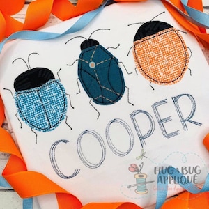 May include: White fabric with three embroidered bugs in blue, teal, and orange, each with a black head. The name "COOPER" is embroidered below the bugs. Orange and blue ribbons frame the image.