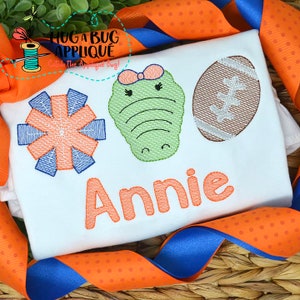 May include: White embroidered t-shirt with a blue and orange pom-pom, a green alligator with a pink bow, and a brown football. The name "Annie" is embroidered in orange below the design.