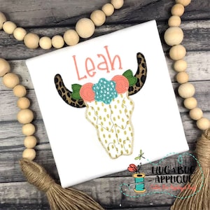 May include: White t-shirt with a gold and white cow skull with pink and turquoise flowers and a leopard print headband. The name "Leah" is embroidered above the skull.