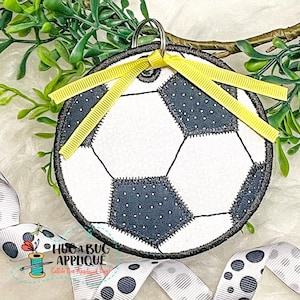 May include: A white and black soccer ball with a yellow ribbon tied around it. The soccer ball is attached to a silver key ring. The soccer ball is on a white background with green leaves.