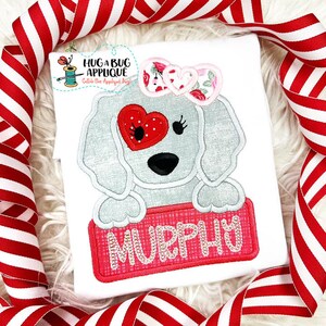 May include: An embroidered grey dog with a red heart over one eye and a pink bow on its head. The dog is holding a pink sign with the name "Murphy" embroidered on it.