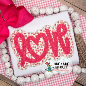 Pode incluir: Camiseta branca com uma aplicação rosa da palavra "love" com um coração no "o". A aplicação tem um padrão floral. O texto "Hug a Bug Applique Catch the Applique Bug!" está abaixo da aplicação.