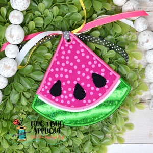 May include: A pink watermelon slice applique with green rind and black seeds. The applique is made of fabric and has a white polka dot pattern.