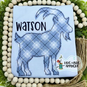 May include: A light blue t-shirt with a blue and white plaid goat applique and the name "Watson" embroidered in black.
