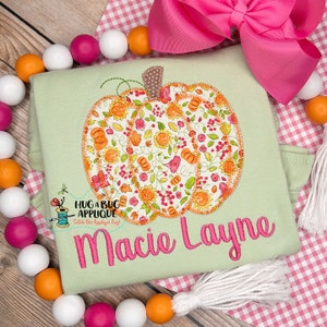 May include: A light green t-shirt featuring a floral pumpkin applique and the name "Macie Layne" embroidered in pink. The shirt is surrounded by a string of white, pink, and orange beads.