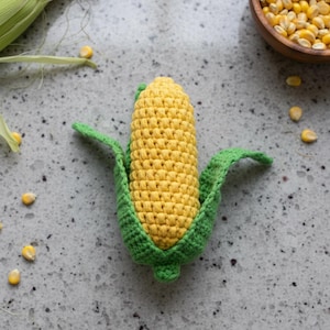 Puede incluir: Un juguete de ganchillo de mazorca de maíz en amarillo y verde. El juguete tiene un diseño detallado con granos visibles. También hay granos de maíz sueltos y un cuenco de granos en la imagen. Un juguete para niños.