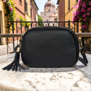 Peut inclure: Un sac bandoulière en cuir noir avec une fermeture éclair dorée et une pampille. Le sac a une forme arrondie et une longue bandoulière réglable. En arrière-plan, une scène de rue européenne.