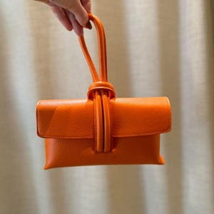 May include: Vibrant orange clutch bag with a top handle. The bag features a flap closure and a textured finish. The handle is knotted at the top. The words "VITA DI LUSSO" are printed in the upper right corner.