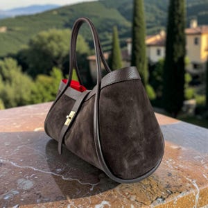 May include: A dark brown suede handbag featuring a structured silhouette, two tall leather handles, and a gold-toned metal turn-lock closure on a central strap. The interior lining is a vibrant red, contrasting with the dark exterior texture.
