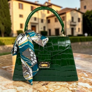 May include: A glossy, emerald green handbag with a crocodile-skin texture. The bag features a top handle and a decorative scarf with a teal, white, and gray pattern. A gold-toned clasp secures the front flap.