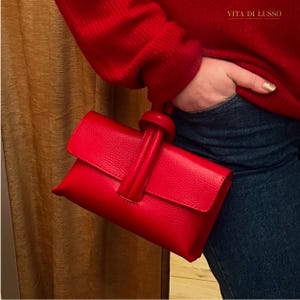 Peut inclure: Une pochette rouge vif avec une finition texturée. La pochette est dotée d'un rabat et d'une poignée avec un nœud décoratif. Le sac est tenu sur un fond de jean bleu et de pull rouge.