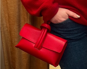 May include: A vibrant red clutch with a textured finish. The clutch features a flap closure and a handle with a decorative knot. The bag is held against a background of blue jeans and a red sweater.