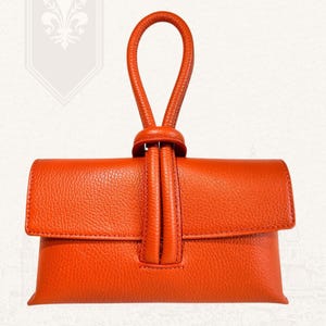 May include: An orange leather clutch with a looped handle. The clutch has a flap closure and a textured surface. The handle is attached to the top of the clutch with a knot detail. The words "VITA DI LUSSO" are printed on the top right.