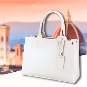 Könnte beinhalten: Eine weiße Ledertasche mit zwei Griffen und einem kleinen Anhänger. Die Tasche hat eine rechteckige Form mit einem strukturierten Design. Der Hintergrund zeigt eine verschwommene Stadtlandschaft mit einer Kuppel.