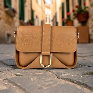May include: A tan-colored crossbody bag with a pebbled texture, featuring a front flap closure secured by a vertical strap and a polished gold-tone metal D-ring hardware accent. The bag has a structured rectangular shape with rounded bottom corners.