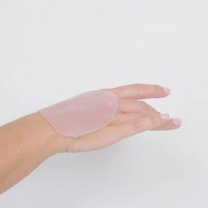 Peut inclure: Un coussinet en silicone rose pâle, de forme ovale, est posé sur une main sur fond blanc. Le coussinet est translucide et semble lisse, probablement pour le confort ou le soutien.