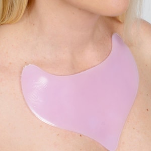 May include: A light pink, crescent-shaped silicone chest patch. The patch is designed to be worn on the chest. The skin is visible around the patch, and the background is a plain white.