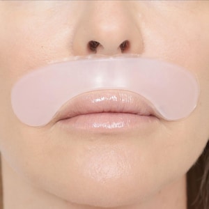 May include: A close-up of a person's face with a light pink lip mask applied over the lips. The mask is a curved shape, covering the upper and lower lips. The skin around the mouth is smooth and the lips appear moisturized.