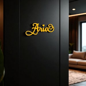 Op de afbeelding: Een gele naamplaat "Aria" in cursiefschrift op een zwarte deur. Het bord heeft een hart boven de letter "i". De achtergrond toont een woonkamer met een bank en een raam.