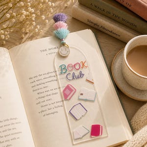 May include: A clear acrylic bookmark featuring the text "BOOK Club" in colorful letters, decorated with small book illustrations. It is topped with a gold-tone smiley face charm and a trio of purple, pink, and light blue fabric tassels.