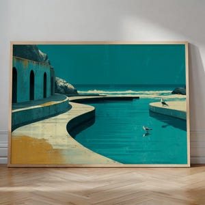 Op de afbeelding: Een ingelijste kunstdruk toont een kustscène met een turquoise waterpartij en een lichtgekleurde betonnen structuur. Twee zeemeeuwen zijn zichtbaar. De lucht is diep turquoise. Het kunstwerk zit in een houten frame.