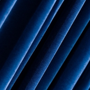 May include: Close-up of deep blue velvet fabric with diagonal folds. The fabric has a soft, textured appearance, with highlights and shadows creating depth. The folds are evenly spaced, suggesting a draped or pleated design.
