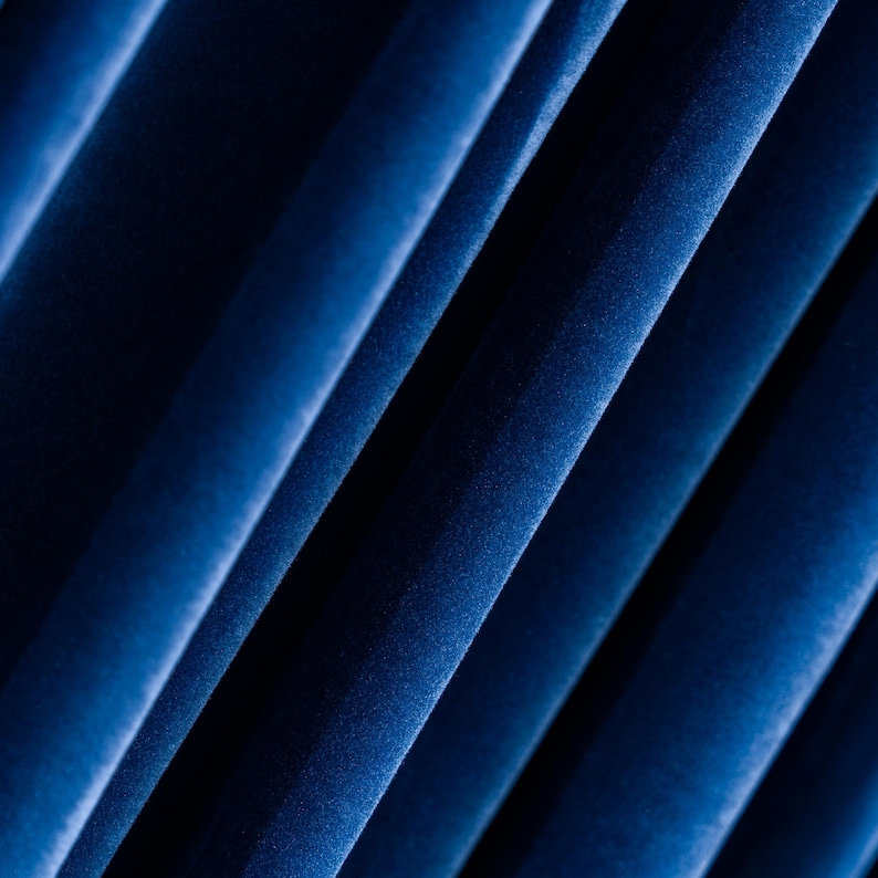 May include: Close-up of deep blue velvet fabric with diagonal folds. The fabric has a soft, textured appearance, with highlights and shadows creating depth. The folds are evenly spaced, suggesting a draped or pleated design.