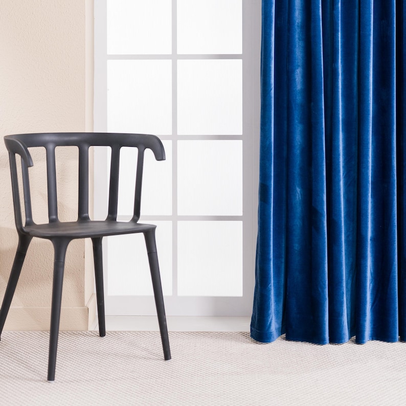 May include: A black plastic chair with a curved back and vertical slats stands near a window with white panes. A vibrant blue velvet curtain hangs to the right. The chair is on a light-colored carpet.