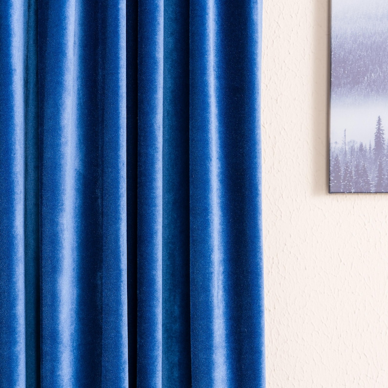 May include: Close-up of a vibrant blue velvet curtain with vertical folds. The fabric has a soft sheen, and the color is a deep, rich shade of blue. The curtain is hanging in front of a light-colored wall.