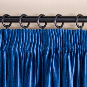 May include: Close-up of a blue velvet curtain with a black curtain rod and rings. The curtain is gathered at the top, creating vertical folds. The fabric has a soft, luxurious appearance, and the color is a deep, rich blue.