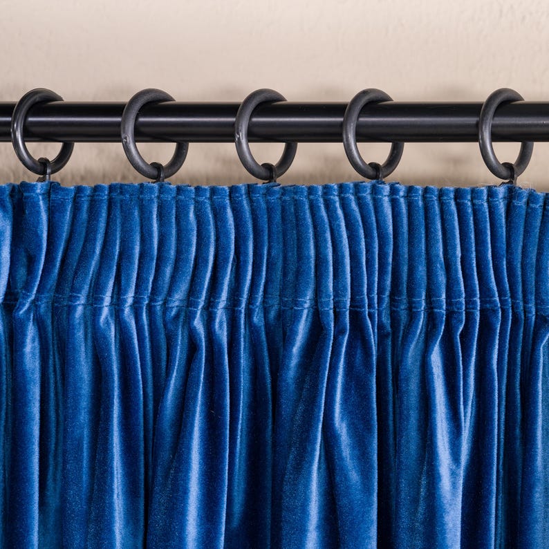May include: Close-up of a blue velvet curtain with a black curtain rod and rings. The curtain is gathered at the top, creating vertical folds. The fabric has a soft, luxurious appearance, and the color is a deep, rich blue.