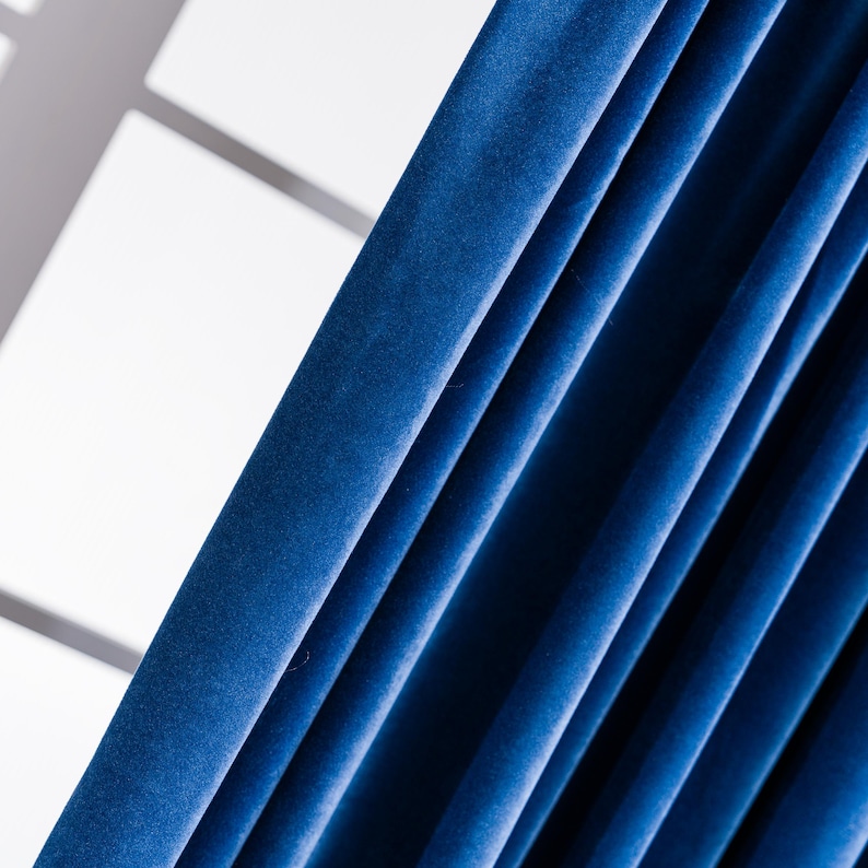 May include: Close-up of deep blue velvet curtains. The fabric is draped in vertical folds, creating shadows and highlights. The curtains are a rich, solid color, and the texture of the velvet is visible. The background is a bright white, with a glimpse of a window.