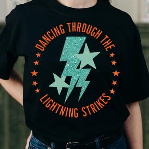 May include: Black t-shirt with a teal glitter lightning bolt and two teal stars. The words "DANCING THROUGH THE LIGHTNING STRIKES" are printed in orange around the design, with orange stars. The shirt is a casual wear item.