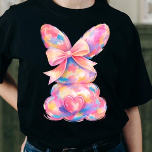 May include: Black t-shirt featuring a colorful, watercolor-style bunny design. The bunny has large ears, a bow, and a heart-shaped detail. The design incorporates shades of pink, blue, yellow, and purple, creating a vibrant and playful aesthetic.