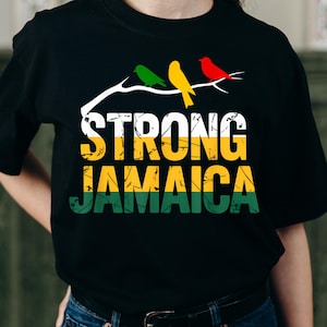 Könnte beinhalten: Schwarzes T-Shirt mit dem Schriftzug "STRONG JAMAICA" in Gelb, Grün und Weiß. Darüber sitzen drei Vögel in Grün, Gelb und Rot auf einem weißen Ast. Das Design feiert Jamaika.