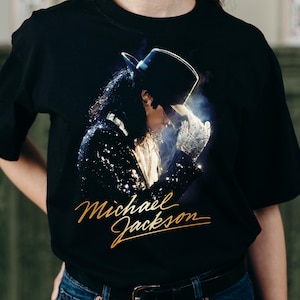 May include: Black t-shirt with a gold "Michael Jackson" script and a portrait of the artist in a black hat and sequined jacket. The image shows a side profile of the singer with a white glove and a smoky effect.