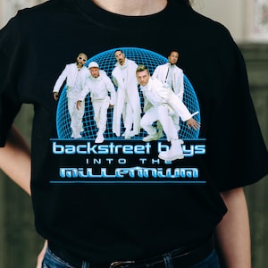 May include: A black t-shirt featuring a graphic of the Backstreet Boys in white outfits against a blue grid sphere. The text reads Backstreet Boys Into The Millennium in a blue font. The design is centered on the front of the garment.