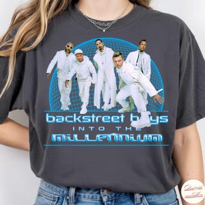 May include: A charcoal gray t-shirt featuring a graphic of the Backstreet Boys in white outfits against a blue grid sphere. The text reads "backstreet boys" and "into the millennium" in a stylized blue font below the group members.