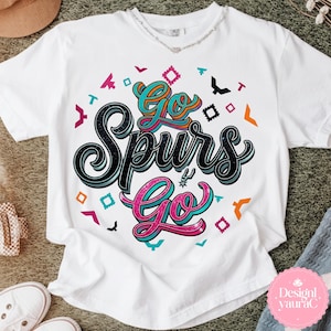 May include: White t-shirt with the text "Go Spurs Go" in a colorful, stylized design. The design includes teal, pink, and black lettering, with decorative shapes in various colors. The shirt is a casual wear item.