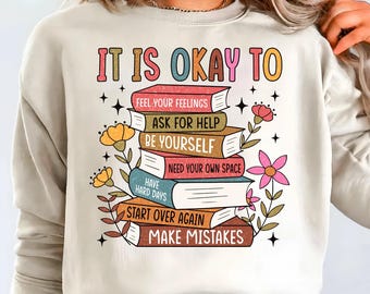 May include: Cream-colored sweatshirt with the text "IT IS OKAY TO" above a stack of books. Each book has a different title, including "FEEL YOUR FEELINGS" and "MAKE MISTAKES." Floral and star graphics surround the books.