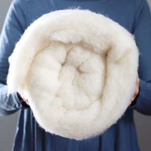 May include: A large, rolled bundle of soft, off-white wool. The wool appears fluffy and dense, with a slightly textured surface. The material is held by a person wearing a blue long-sleeved shirt. This wool is likely used for crafts or textile projects.