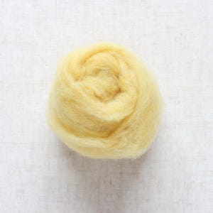 May include: A close-up of a ball of yellow wool roving. The wool is tightly wound, displaying a spiral pattern. The colour is a soft, pale yellow, and the texture appears fluffy and soft. The background is a white, textured surface.