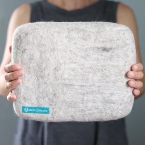 May include: A large, light gray, rectangular cushion made of felted wool. The cushion has a small, teal blue label with the text "GREY FOX FELTING" on it.