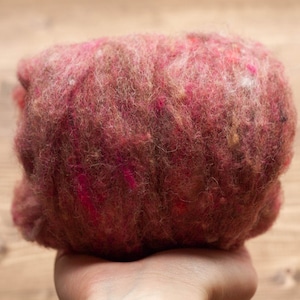 May include: A close-up of a ball of pink, brown, and white wool yarn. The yarn is soft and fluffy, and it has a speckled appearance.