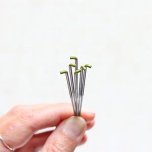 May include: A small bundle of silver-colored floral wire with green, L-shaped tips. The wires are held in a person's hand, with a plain white background.