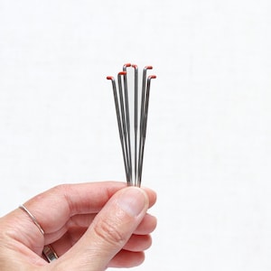 May include: A bundle of thin, silver metal knitting needles with red tips. The needles are held in a hand against a white background. The needles are straight with a slight curve at the end.