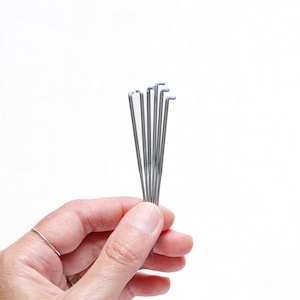 May include: A hand holds a bundle of silver knitting needles with blue tips against a white background. The needles are thin and straight, with a slight bend at the top. The needles are held together, fanned out slightly.