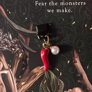 May include: A black clip with a gold-colored loop holding a red chili pepper charm and a white pearl. The text "Fear the monsters we make" is above the charm.